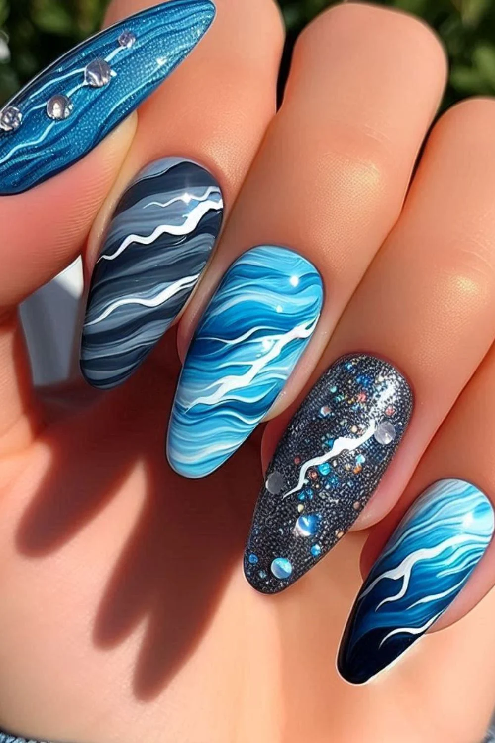 A set of long, almond-shaped nails featuring a blue and white wave design. Some nails have a glossy marbled effect, while one has a black glittery base with small crystal embellishments, resembling ocean waves and shimmering water.