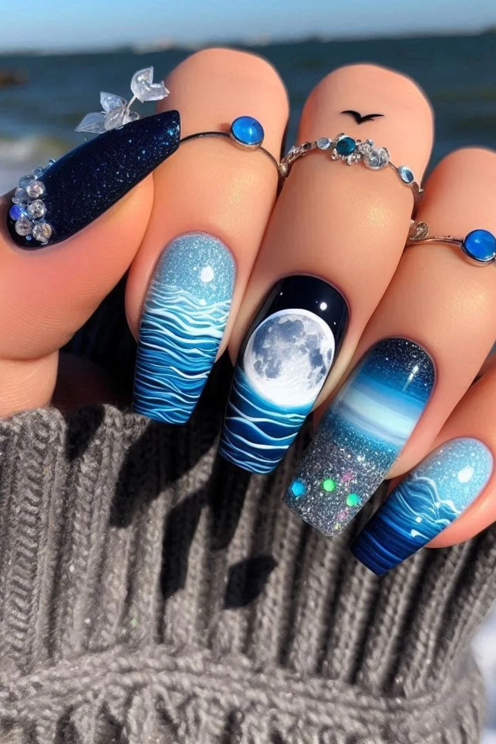 A set of long, coffin-shaped nails with a midnight ocean theme. The nails feature a glowing full moon over rippling ocean waves, a starry night sky, glittery water reflections, and 3D embellishments, evoking a magical seaside night.