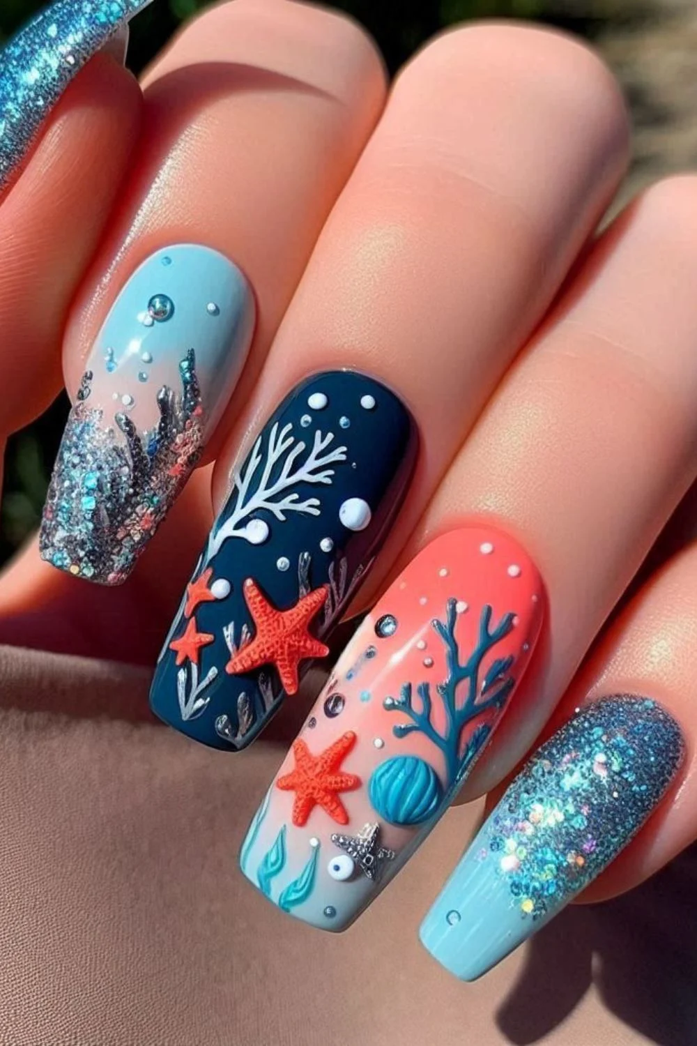 A set of long, coffin-shaped nails with a coral reef theme. The nails feature hand-painted coral branches, 3D starfish, seashells, glittery ocean water, and tiny bubble embellishments, creating a detailed underwater-inspired look.