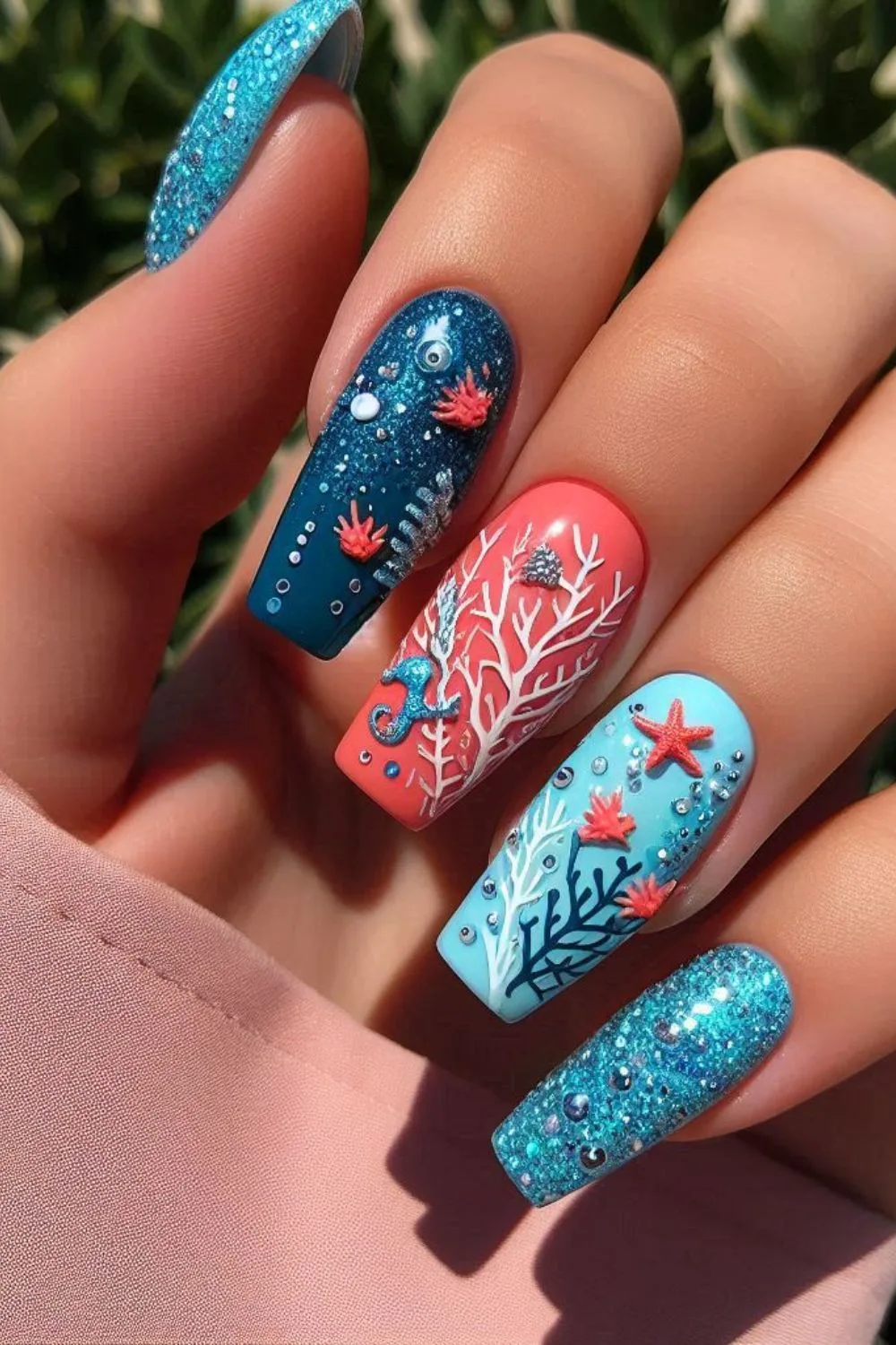 A set of long, coffin-shaped nails with a coral reef theme. The nails feature hand-painted coral branches, a 3D seahorse, starfish, and sparkling ocean textures in shades of deep blue, coral pink, and aqua, evoking an enchanted underwater scene.
