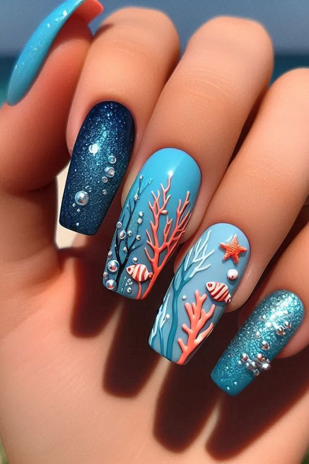 A set of long, coffin-shaped nails featuring an underwater theme. The nails showcase hand-painted coral in soft pink and white, tiny 3D fish, starfish, and bubble embellishments, with shimmering blue gradients mimicking the ocean depths.