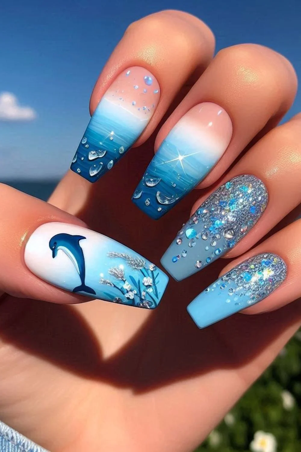 A set of long, coffin-shaped nails featuring an ocean-inspired design. The nails have a blue-to-white ombre effect, a hand-painted dolphin, sparkling water droplet details, and glittery embellishments, mimicking the calm and beauty of the sea.