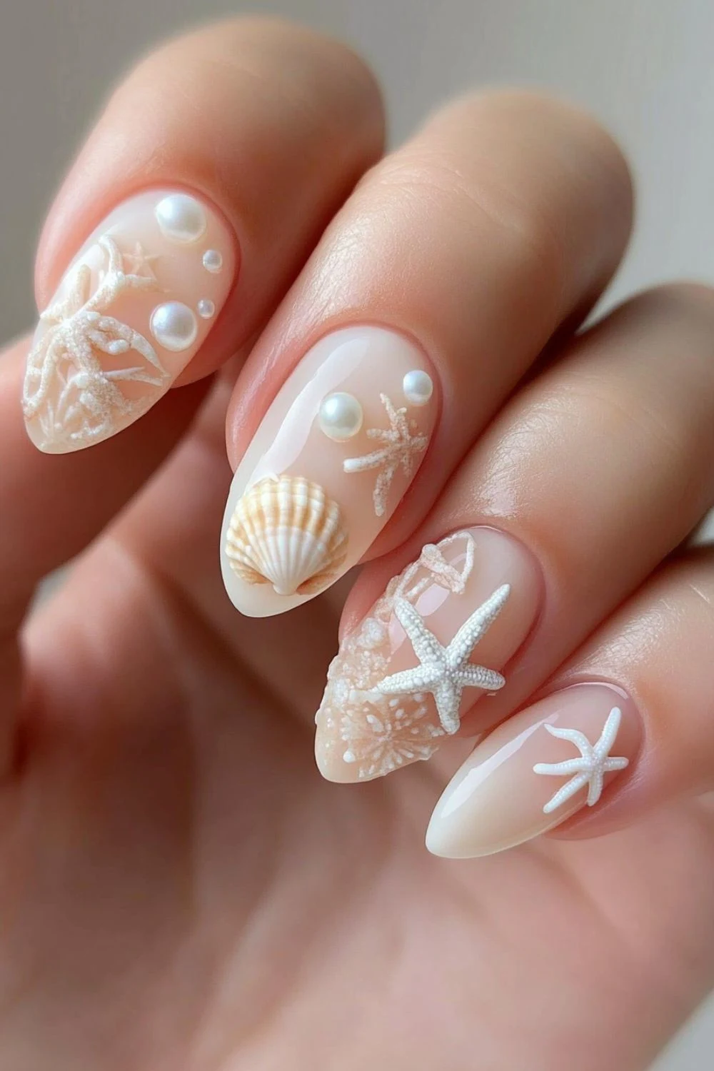 A set of almond-shaped nails featuring an elegant beach-inspired design. The nails have a sheer nude base with 3D seashells, starfish, pearls, and textured sand-like accents, evoking a serene coastal aesthetic.