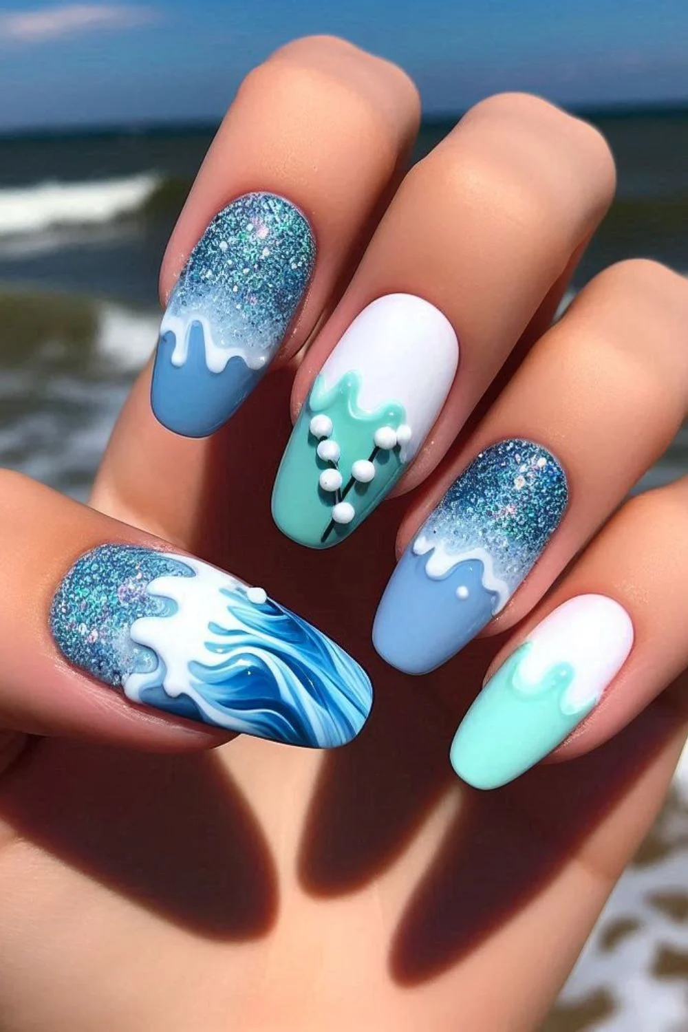 A set of medium-length almond-shaped nails featuring a sea-inspired design with blue and turquoise tones, glitter, white wave patterns, and 3D pearl details, resembling ocean waves and foam.