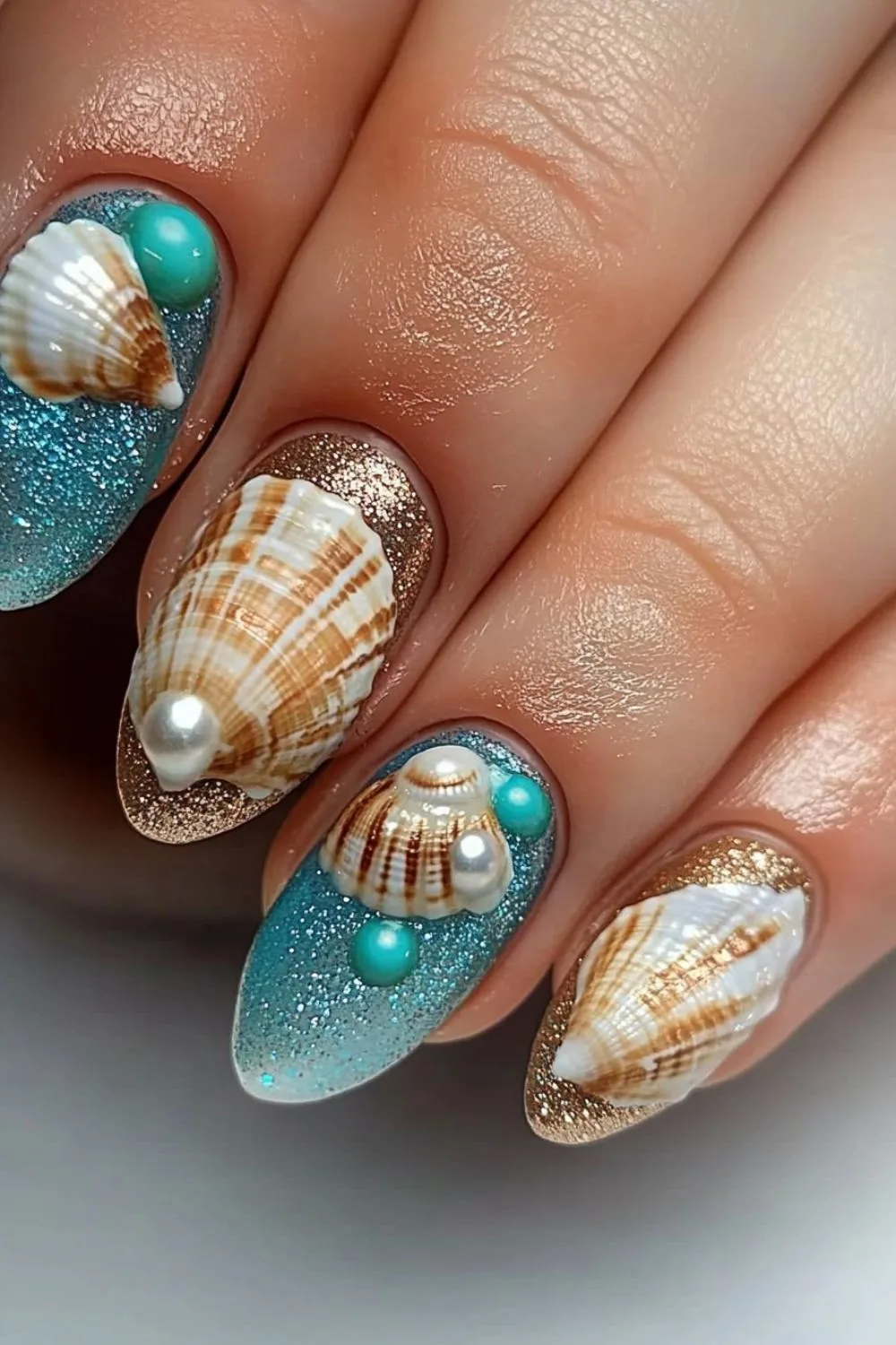 A close-up image of sea jewel-inspired nail art. The nails feature intricate designs with seashells, pearls, and turquoise beads, adorned with glittery blue and gold polish that creates a shimmering oceanic effect.