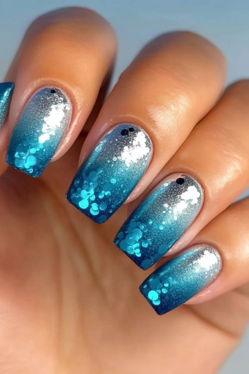 A close-up image of nails with a gradient design transitioning from silver at the base to vibrant blue at the tips, adorned with glitter and small circular confetti decorations in shades of blue and white.