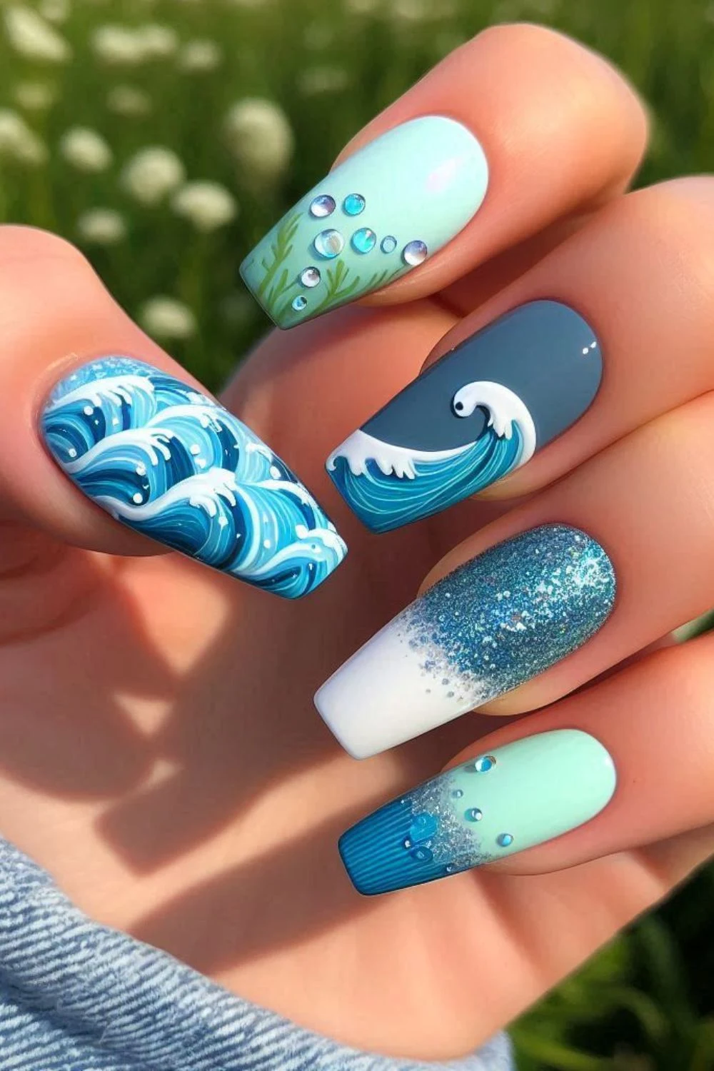 A set of long, coffin-shaped nails with a sea-inspired design. The nails display hand-painted ocean waves, a stylized crashing wave, glittery blue ombre, and light blue nails adorned with tiny bubble-like embellishments.