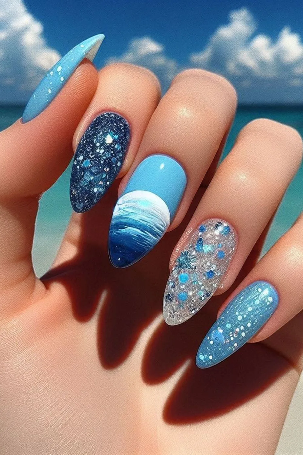 Almond-shaped nails with ocean wave art, deep blue glitter, and transparent accents featuring blue and silver embellishments.