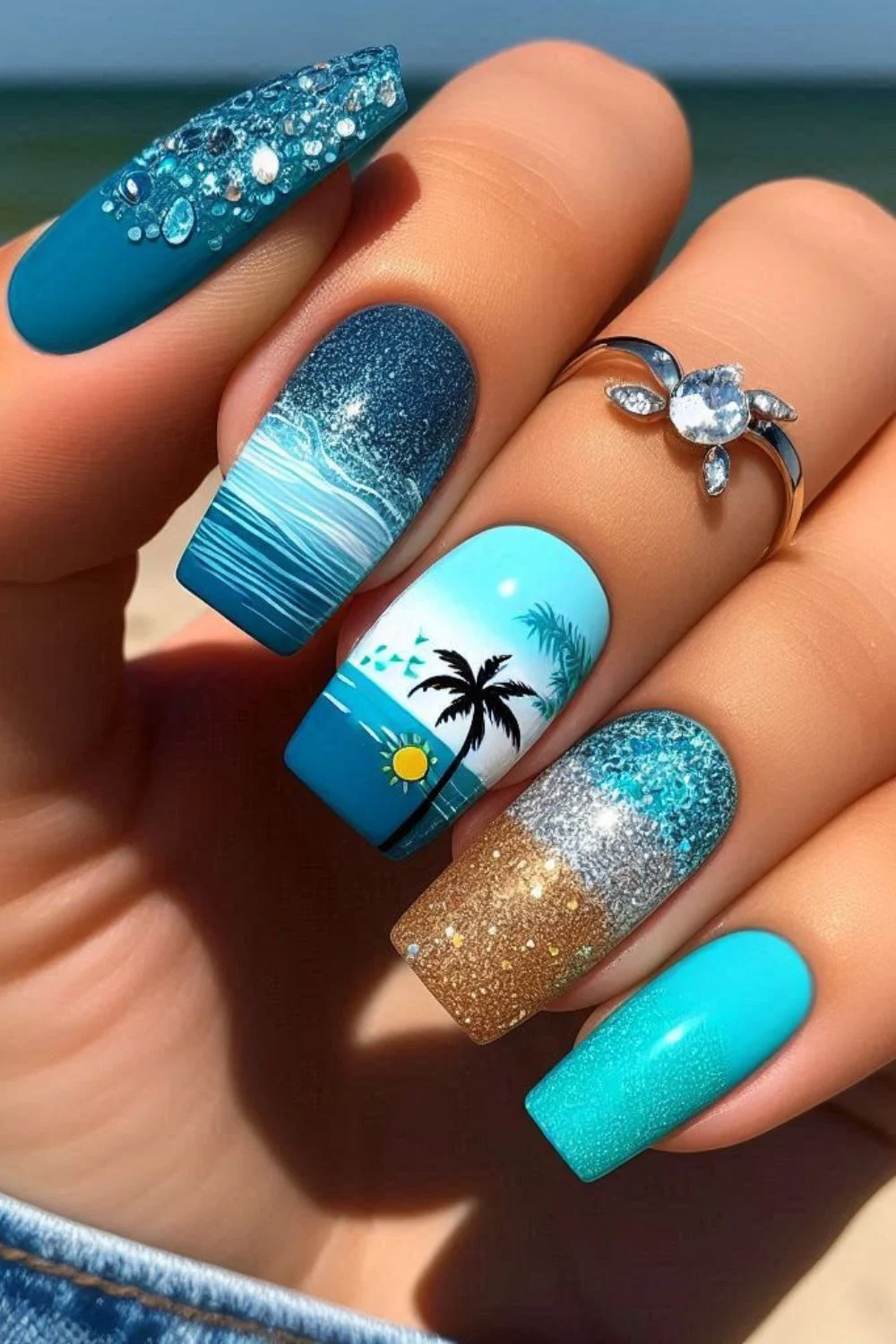 Square-shaped nails featuring palm tree nail art, ocean waves, gold glitter sand, and blue rhinestone accents for a tropical beach design.