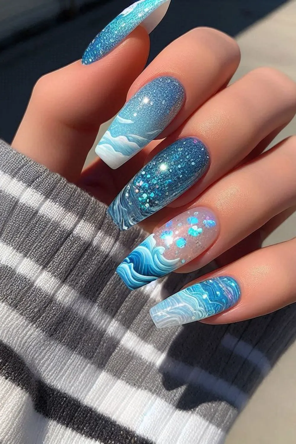 Long square nails with blue and white ocean waves, glitter, and iridescent accents for a sea-inspired manicure.