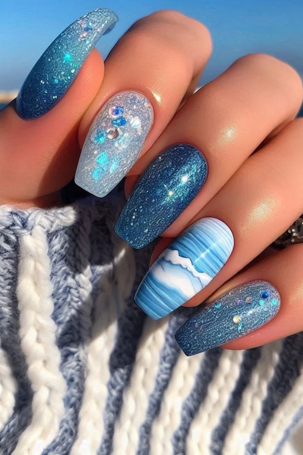 Long square nails with deep blue shimmer, white wave patterns, and iridescent glitter for a sparkling ocean-inspired design.