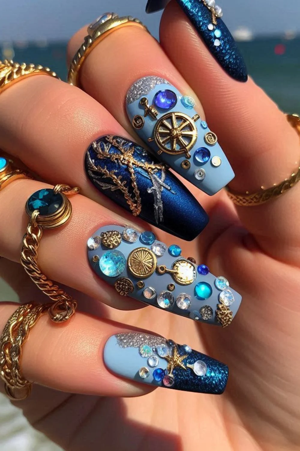 Long blue and gold nails with ship wheel charms, anchors, and gemstones for a deep-sea-inspired treasure manicure.