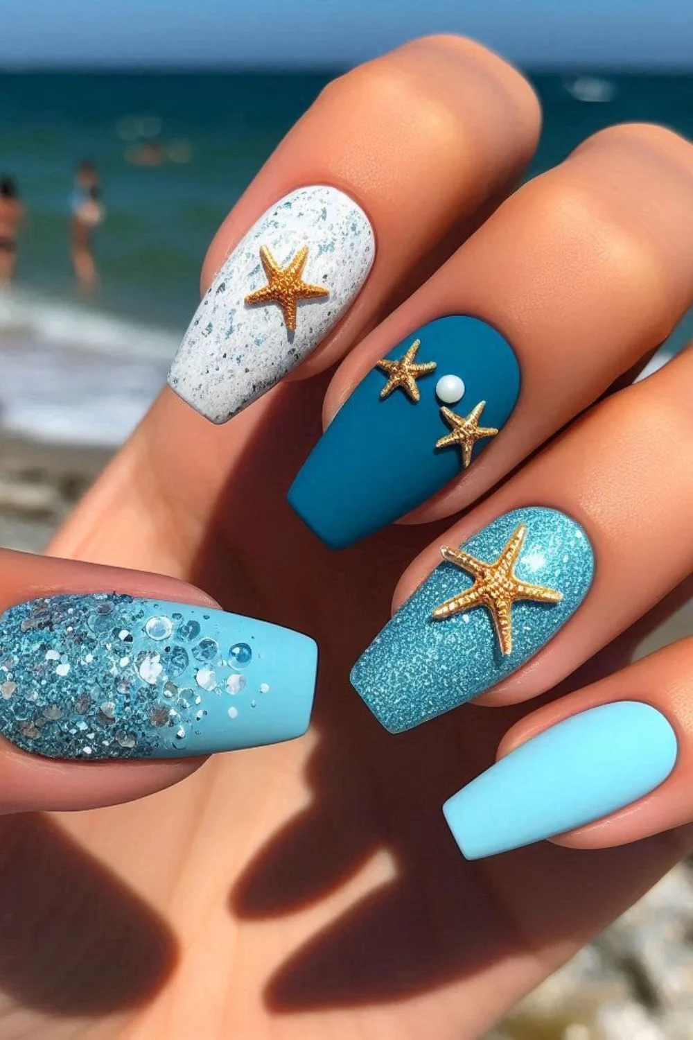Blue and white nails with 3D golden starfish and pearl accents, featuring glitter and textured designs for a beach-inspired manicure.