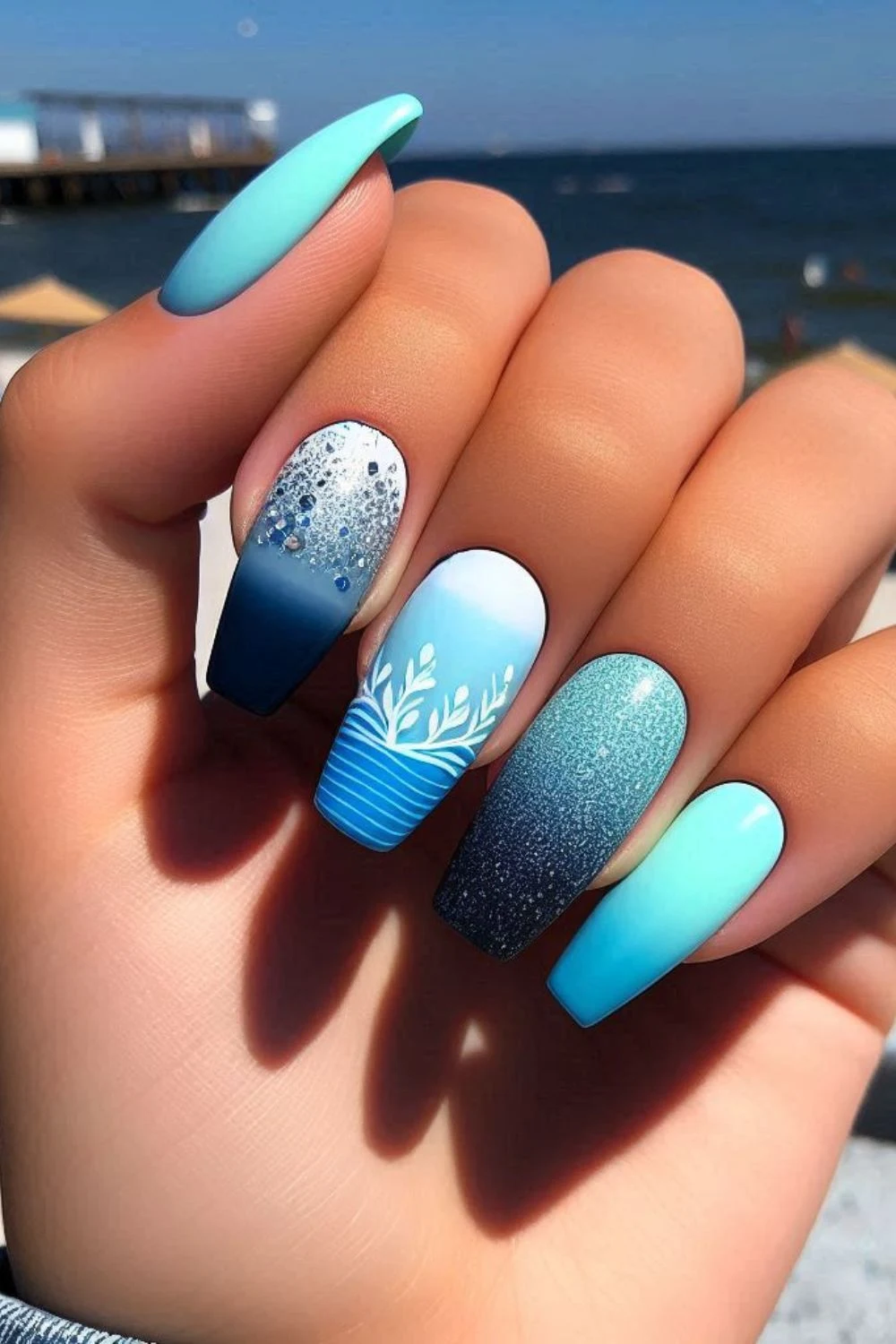 Blue gradient ocean-themed nails with white botanical accents, glitter, and ombré effects, evoking the beauty of the sea.