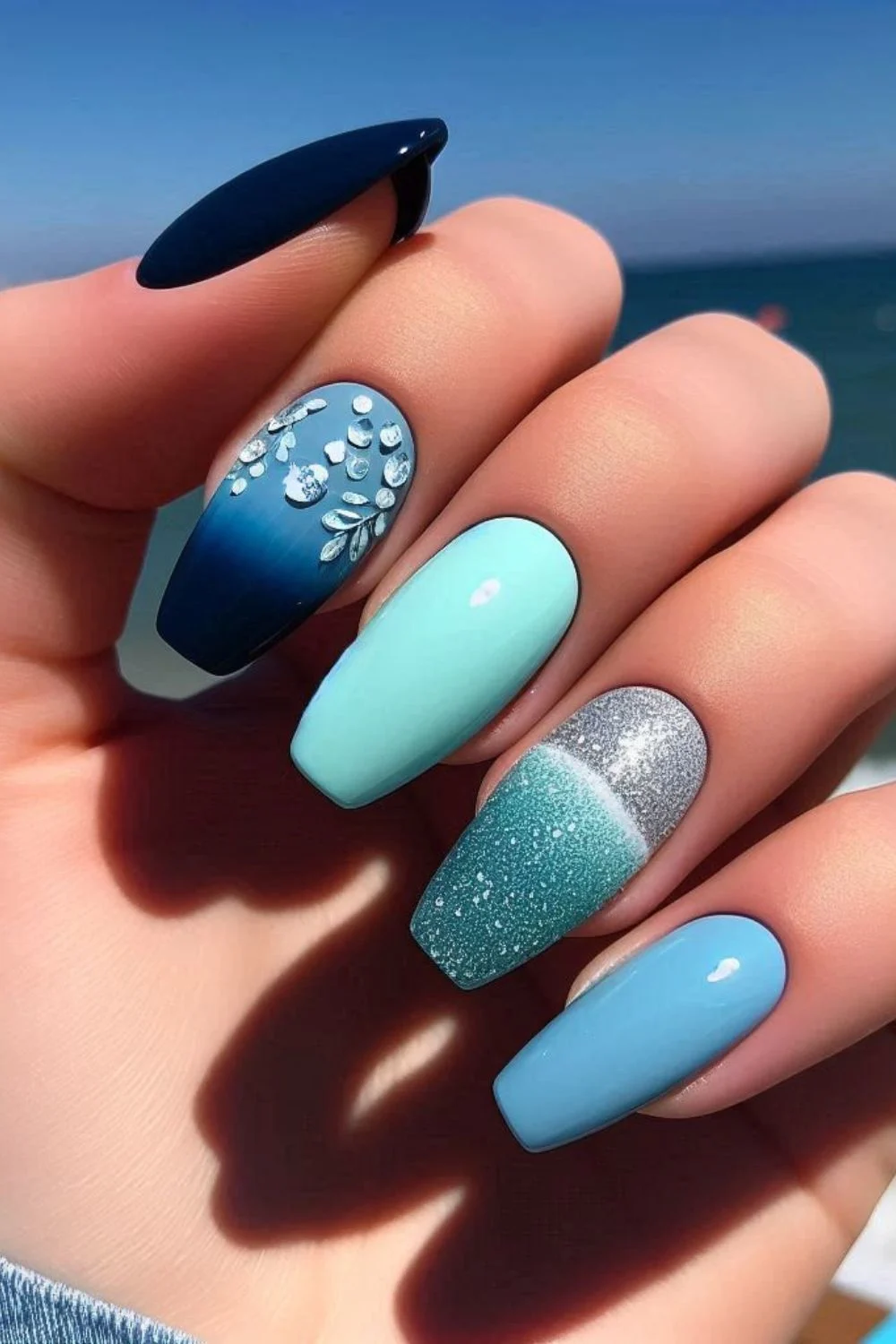 A close-up of a hand with sea-inspired nail art featuring ombre blue shades, glitter, and crystal embellishments, set against a blurred ocean background.