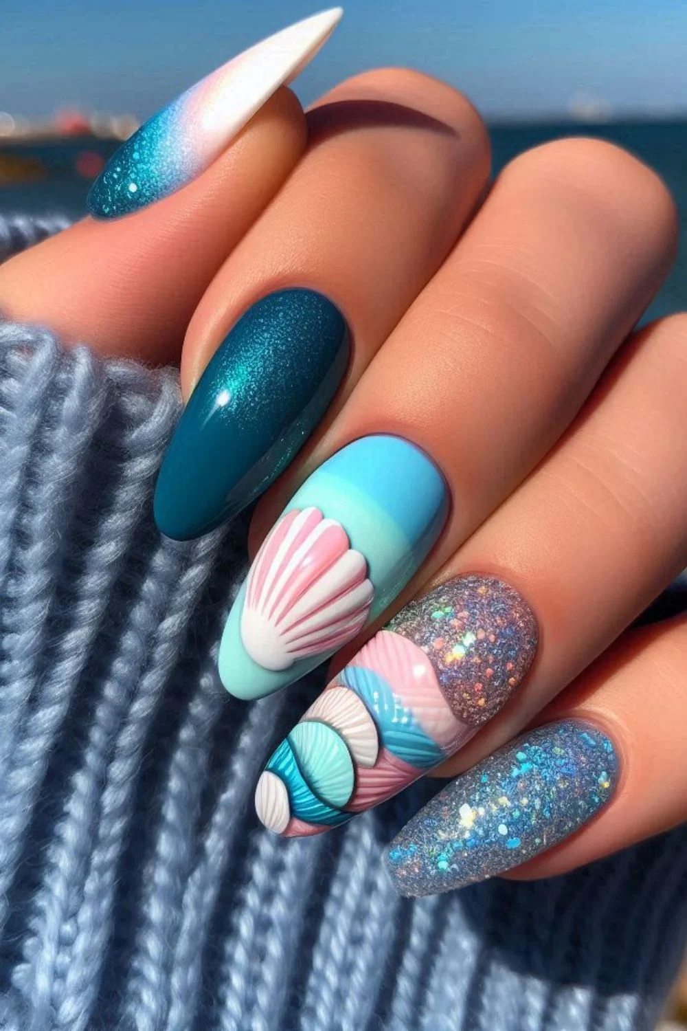 A set of long, almond-shaped nails with a seashell-inspired design. The nails feature 3D pastel pink, blue, and white seashells, glittery accents, an ombre effect, and a deep teal shimmer, creating a beachy, oceanic look.