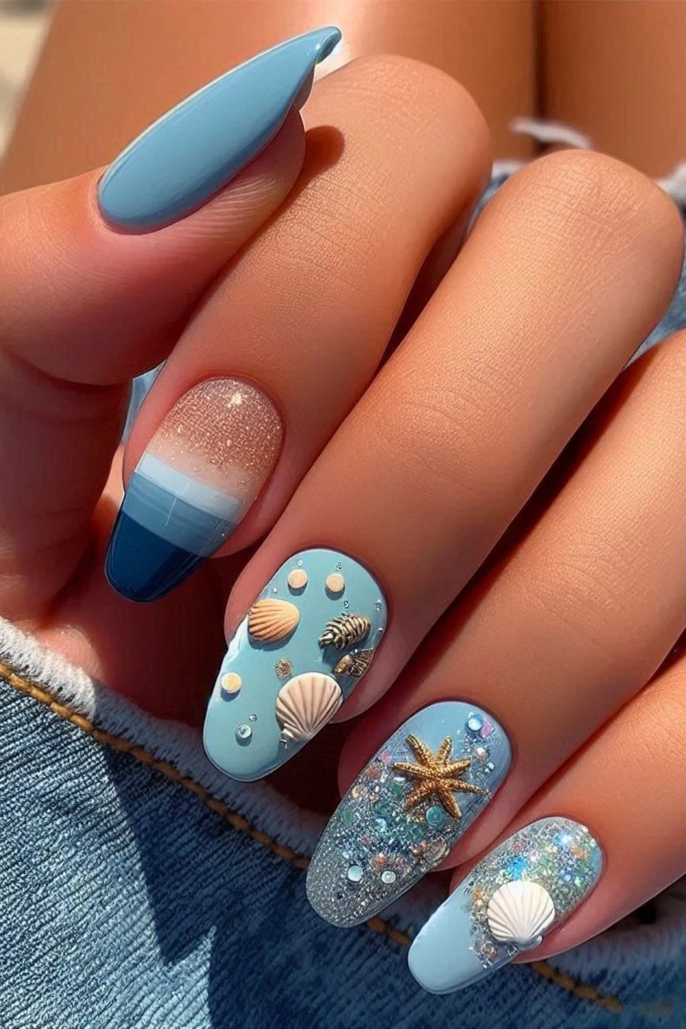 A close-up of a hand with beach-themed nail art featuring blue ombre, glitter, and 3D seashell and starfish embellishments, set against a denim background.