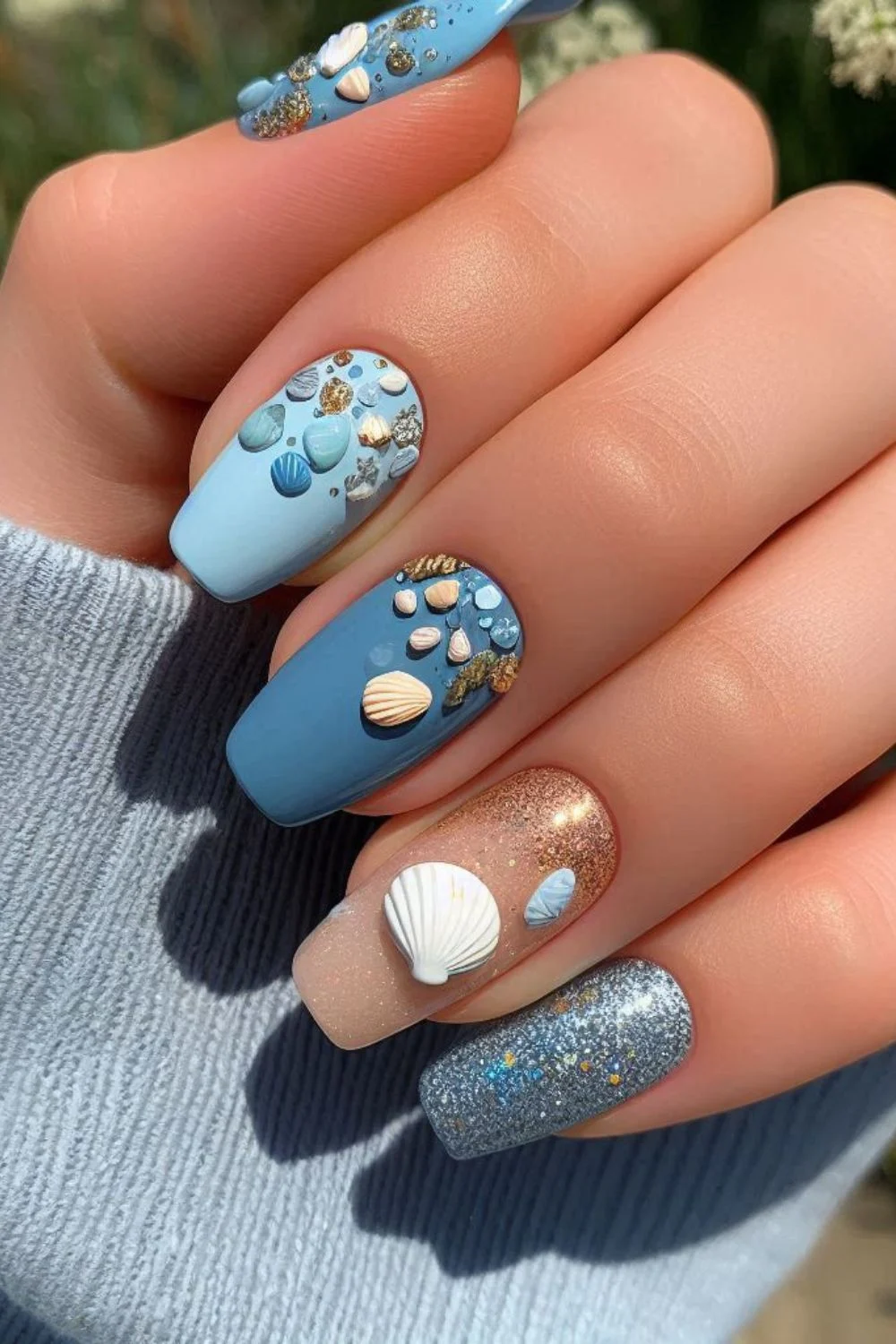 A close-up of a hand showcasing sea-themed nails with a mix of soft blue and nude tones, adorned with 3D seashells, glitter, and small gold embellishments. The nails have a combination of matte and glossy finishes, creating a luxurious ocean-inspired look.
