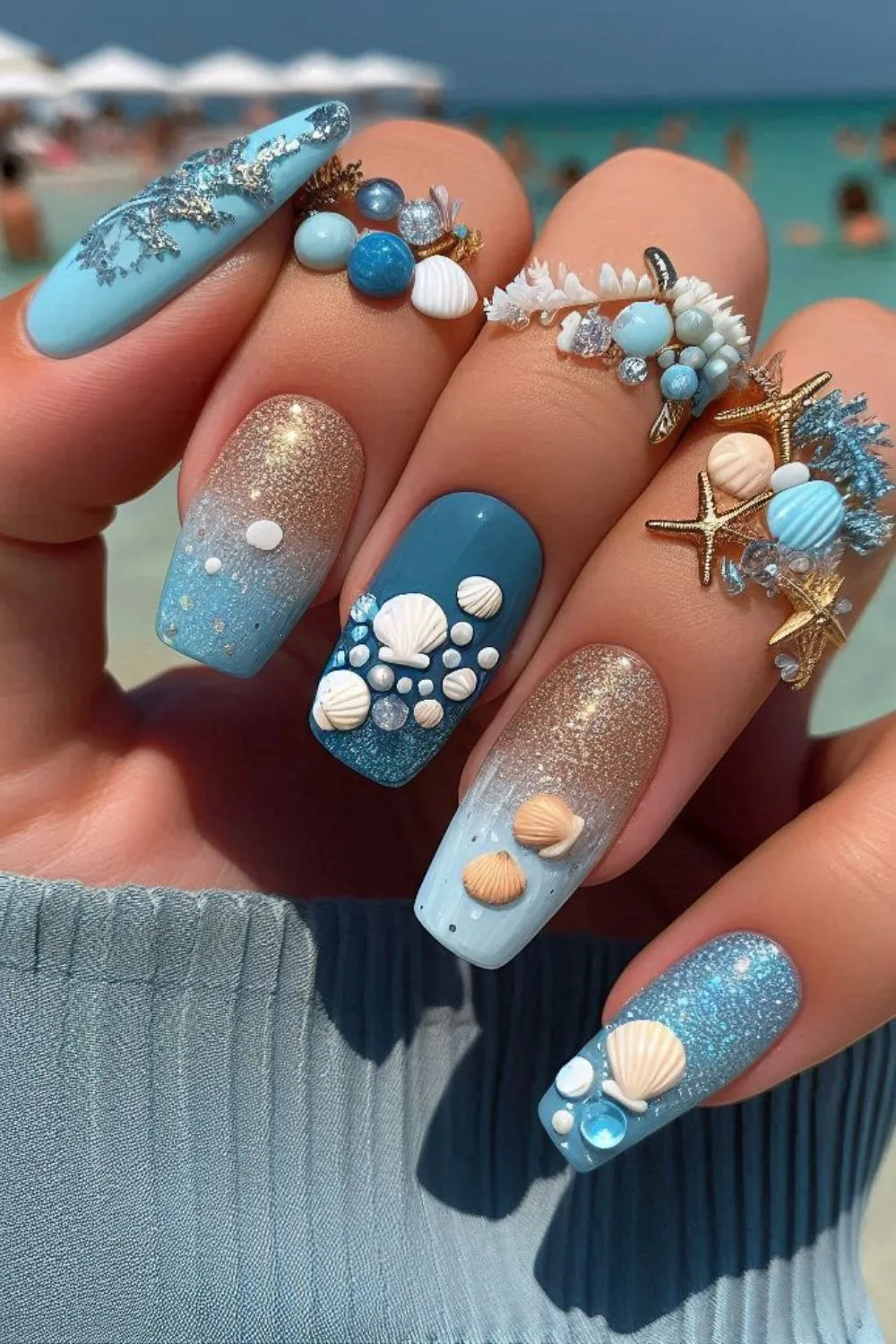 A hand with ocean-inspired nails featuring seashells, glitter, and blue gradients, paired with nautical jewelry.