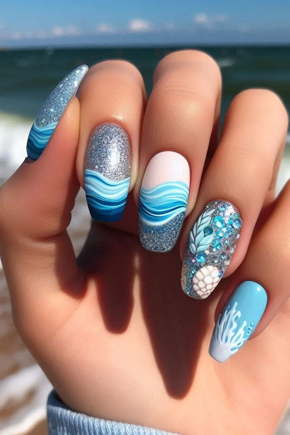 A close-up of a hand with ocean-inspired nails, showcasing blue wave patterns, glitter, and seashell accents against a beach background.