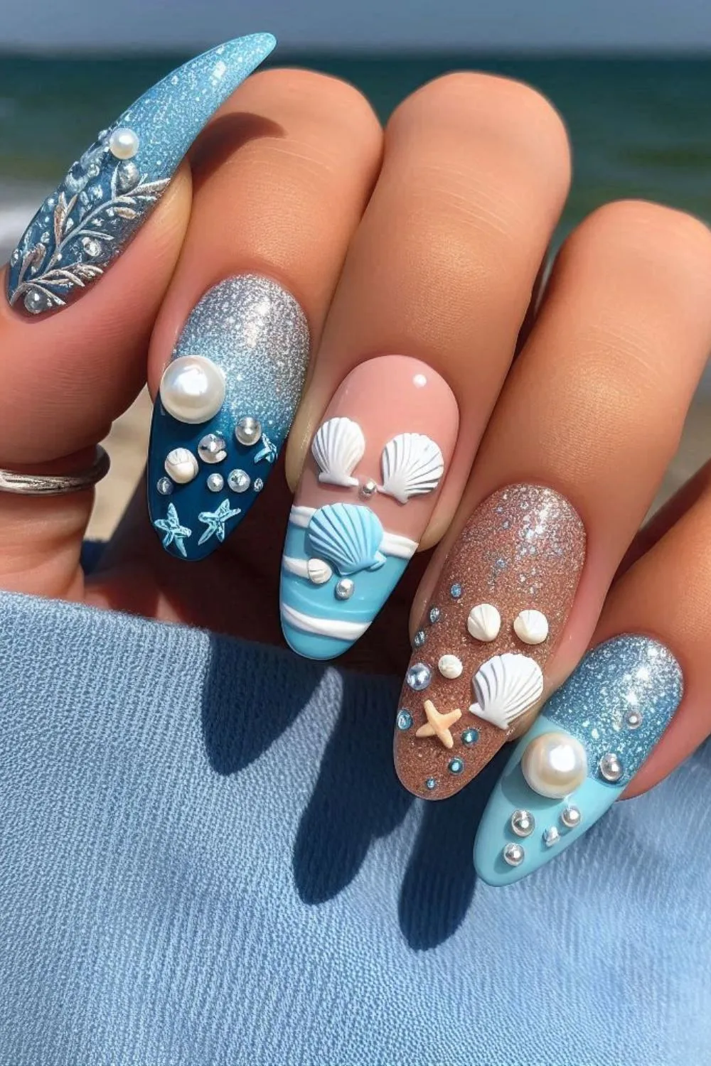 A close-up of beach-inspired nails with seashell accents, pearls, and glittery blue and sand-colored polish against a beach background.