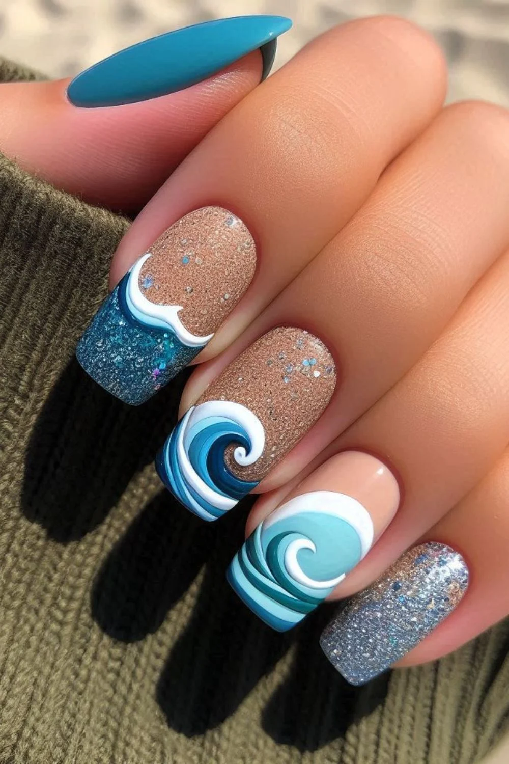 A set of long, coffin-shaped nails featuring a beach-inspired design. The nails have a mix of glittery sand-colored polish, deep blue waves with white foam accents, and shimmering silver details, mimicking ocean waves washing onto the shore.