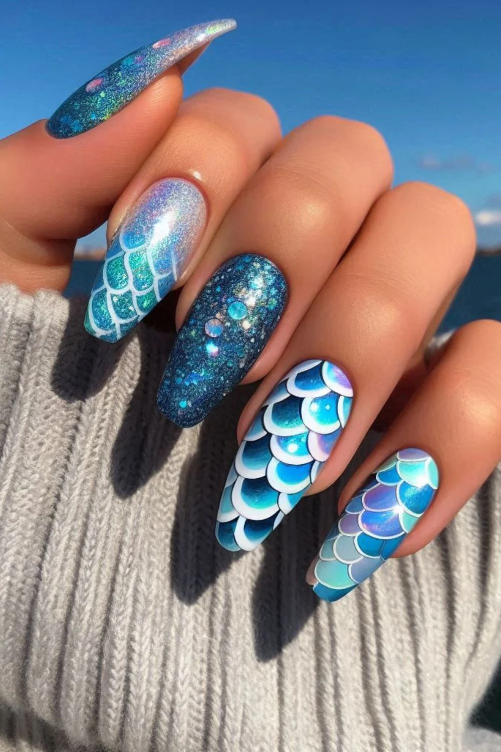 Close-up of a hand with mermaid-themed nails, showcasing iridescent blue scales, glitter, and shimmering ocean tones.