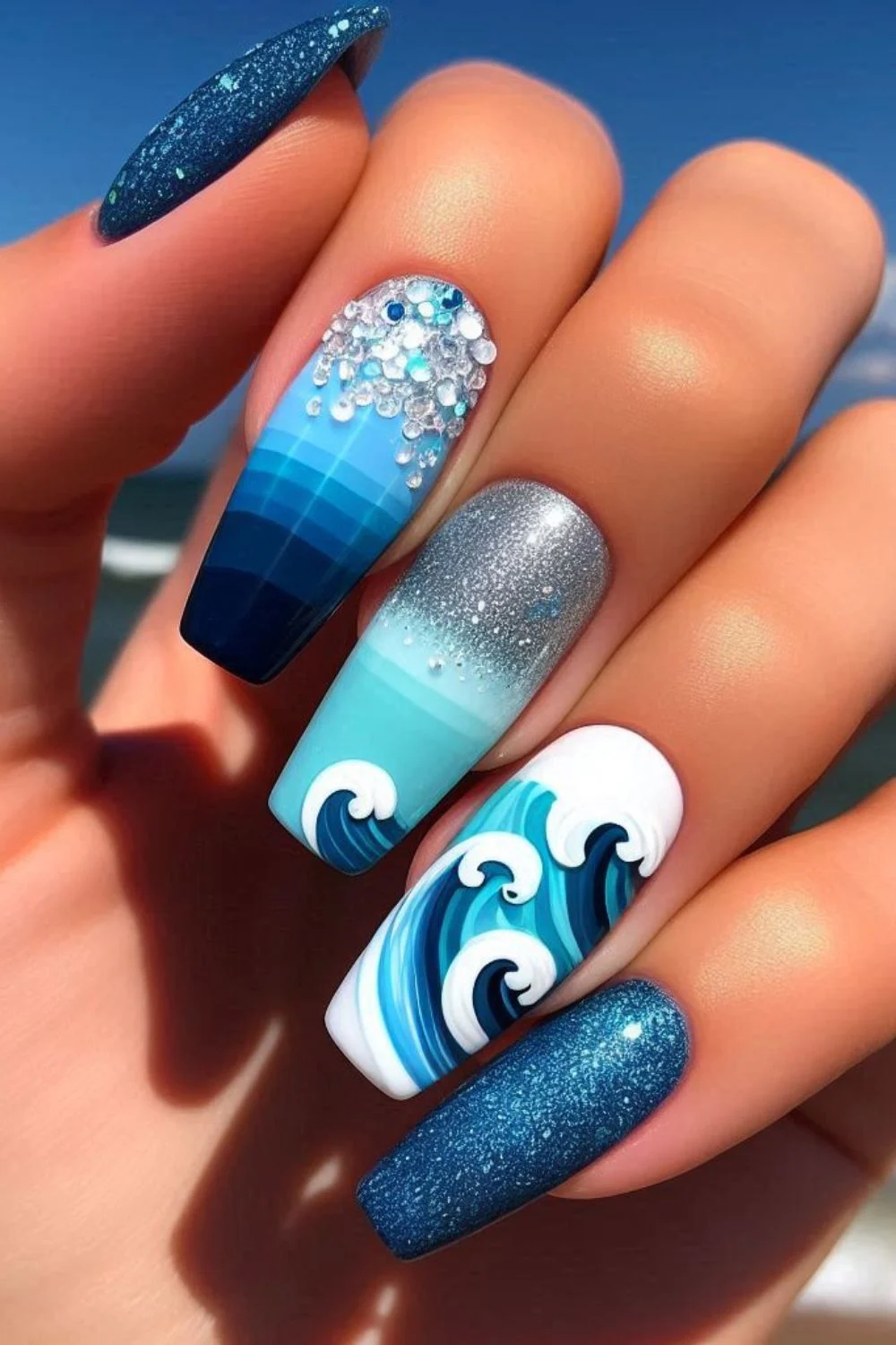 Close-up of ocean-themed nails with blue wave patterns, glitter accents, and crystal details against a beach backdrop.