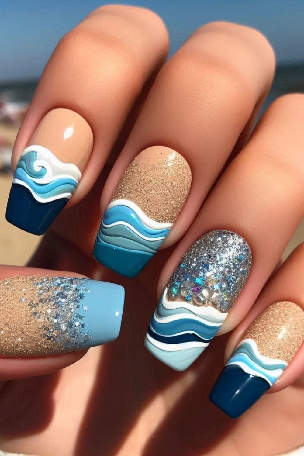 A set of long, coffin-shaped nails featuring a beach-inspired design. The nails have glittery sand-colored polish, layered blue and white waves, and sparkling embellishments, resembling waves crashing onto a sandy shore.