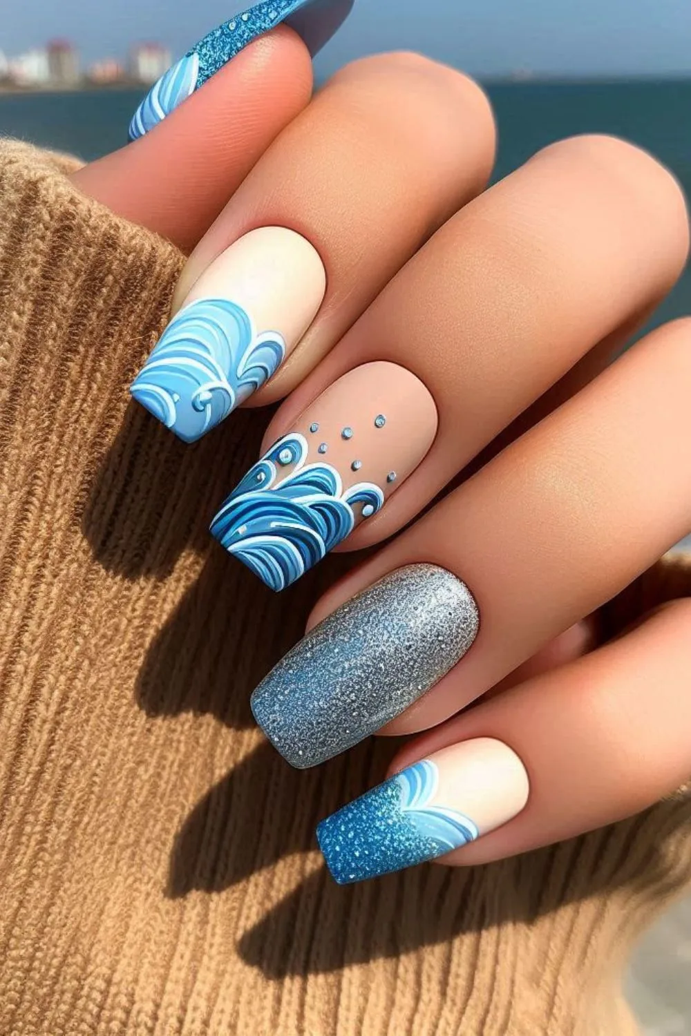 A set of long, coffin-shaped nails featuring a soft wave design. The nails include hand-painted blue ocean waves, nude and beige bases, shimmering silver polish, and tiny water droplet embellishments, evoking a calm and elegant seaside aesthetic.
