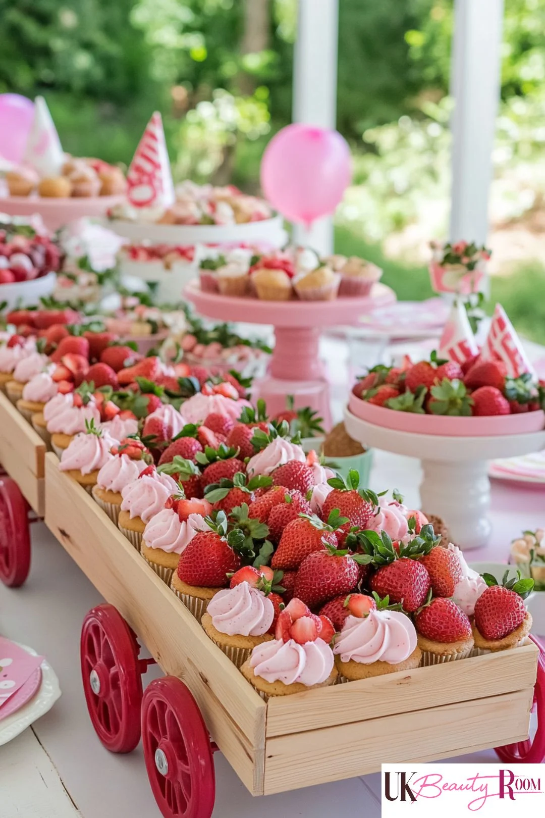A wooden wagon centerpiece filled with strawberry-topped cupcakes and fresh strawberries, set on a pink-themed outdoor party table with balloons and desserts.